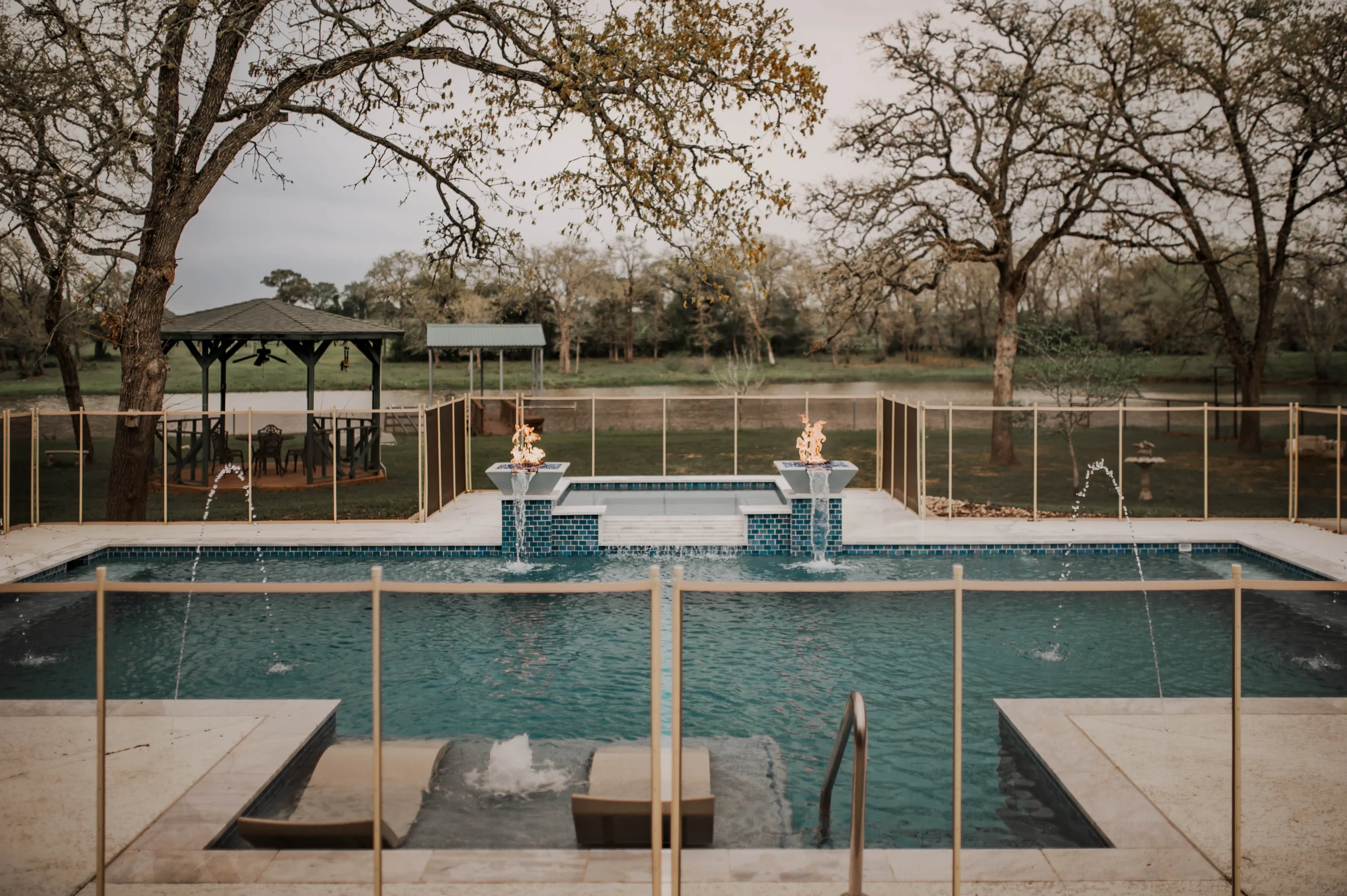 A luxurious backyard pool features water fountains and flame accents, surrounded by trees and a gazebo