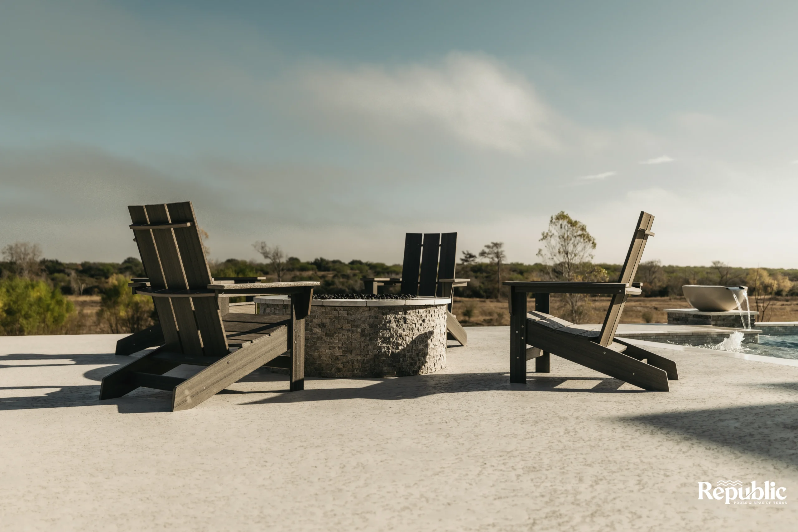 A serene outdoor space featuring wooden lounge chairs facing a stone fire pit, with a lush landscape and clear sky in the background