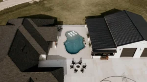 Aerial view of a modern home with a pool, outdoor seating area, and a black-roofed garage on a grassy lot