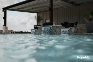A serene pool scene featuring cascading waterfalls, a modern patio, and a distant house under cloudy skies