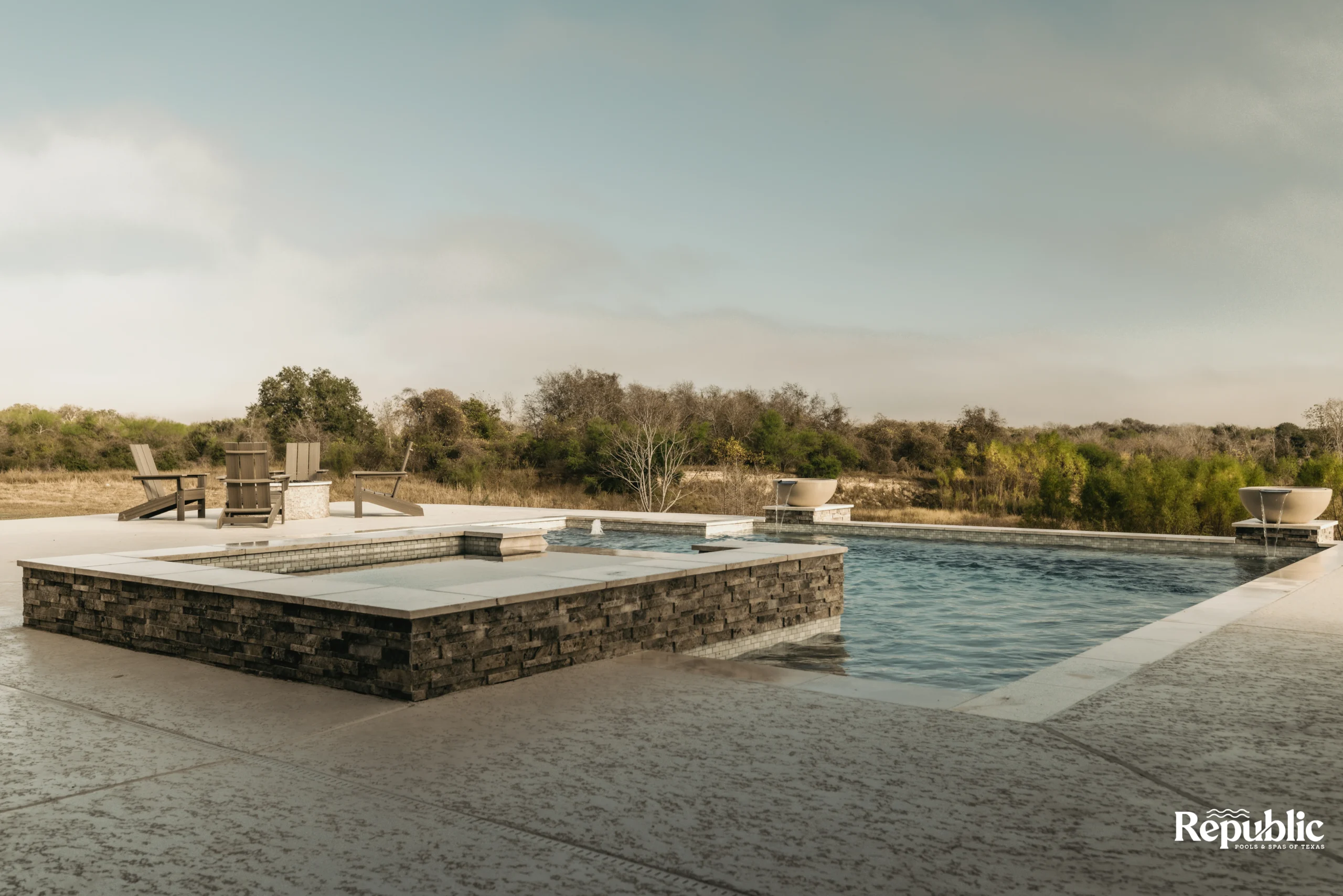 Serene outdoor pool area featuring a stone hot tub, lounge chairs, and a backdrop of lush greenery under a soft sky